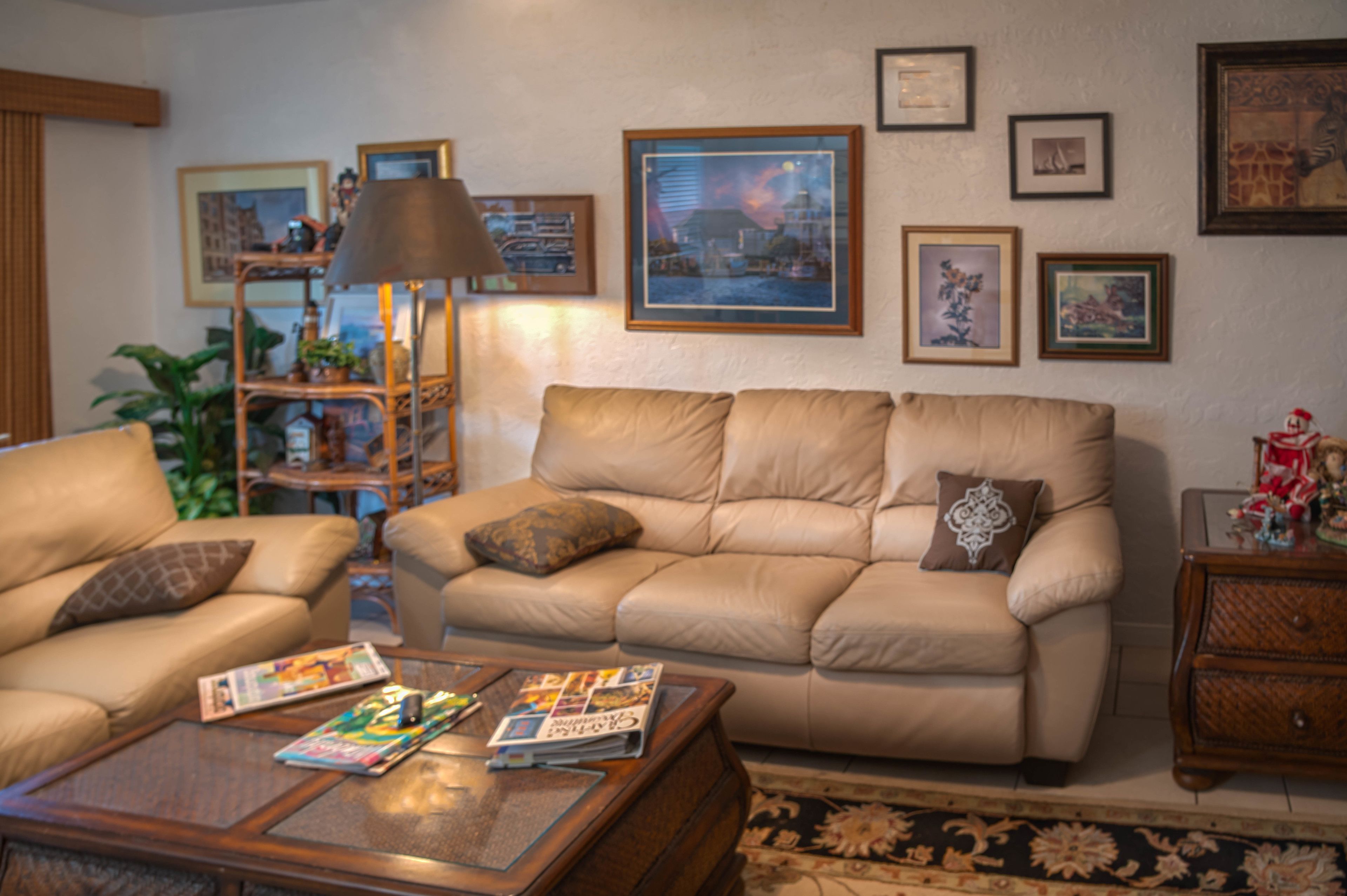 A living room with a beige sofa, a coffee table with magazines, a lamp, and framed pictures on the wall.