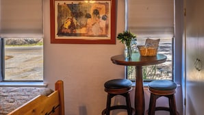 Une pièce comprenant un lit, une petite table avec deux tabourets, un tableau abstrait encadré et une fenêtre avec des stores.