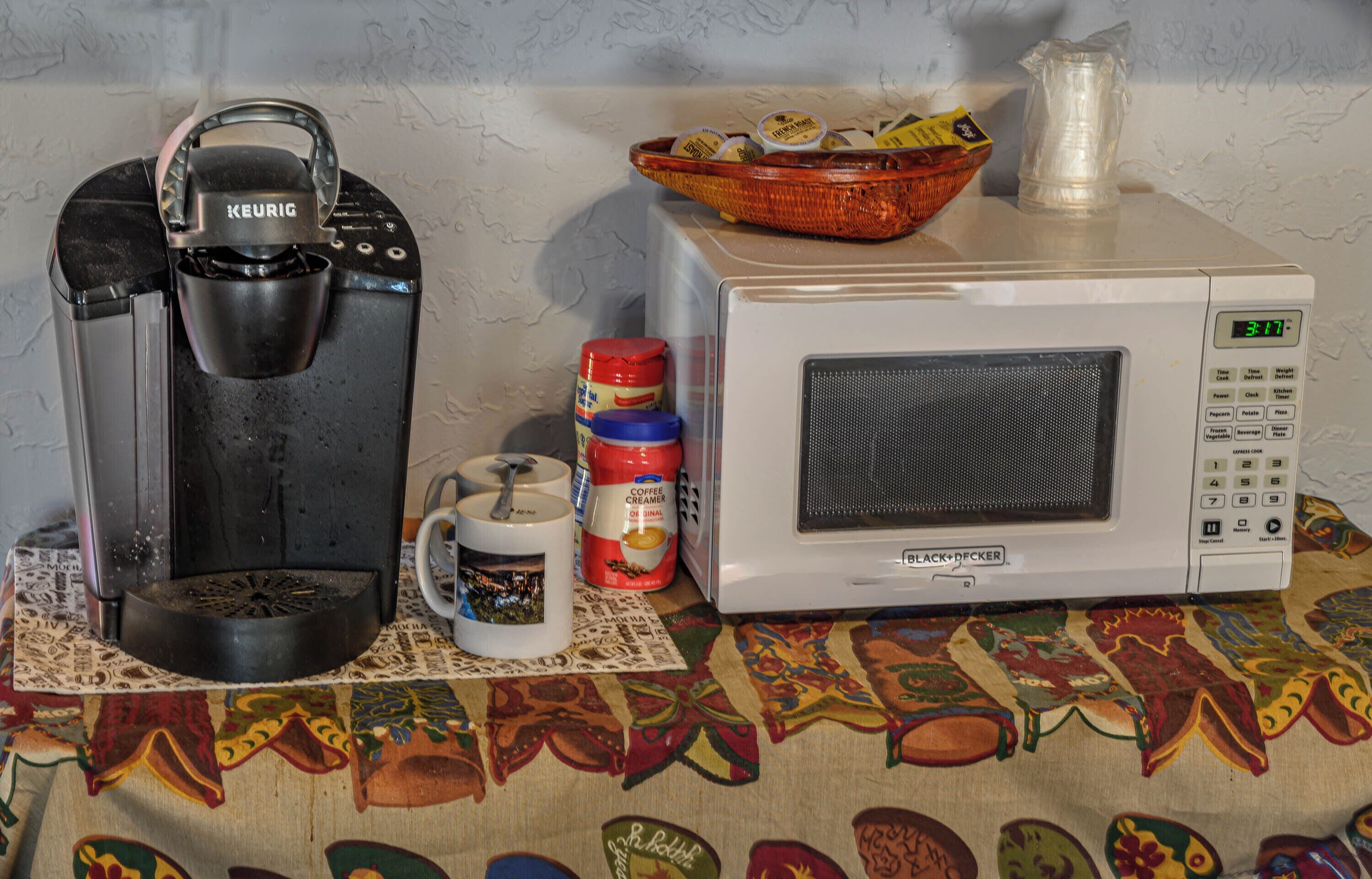 A coffee maker, a mug, a microwave, and a basket of food containers on a table with a patterned tablecloth.