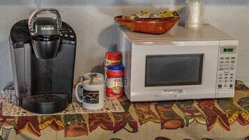 En kaffemaskine, en kop, en mikrobølgeovn og en kurv med madbeholdere på et bord med et mønstret borddække.