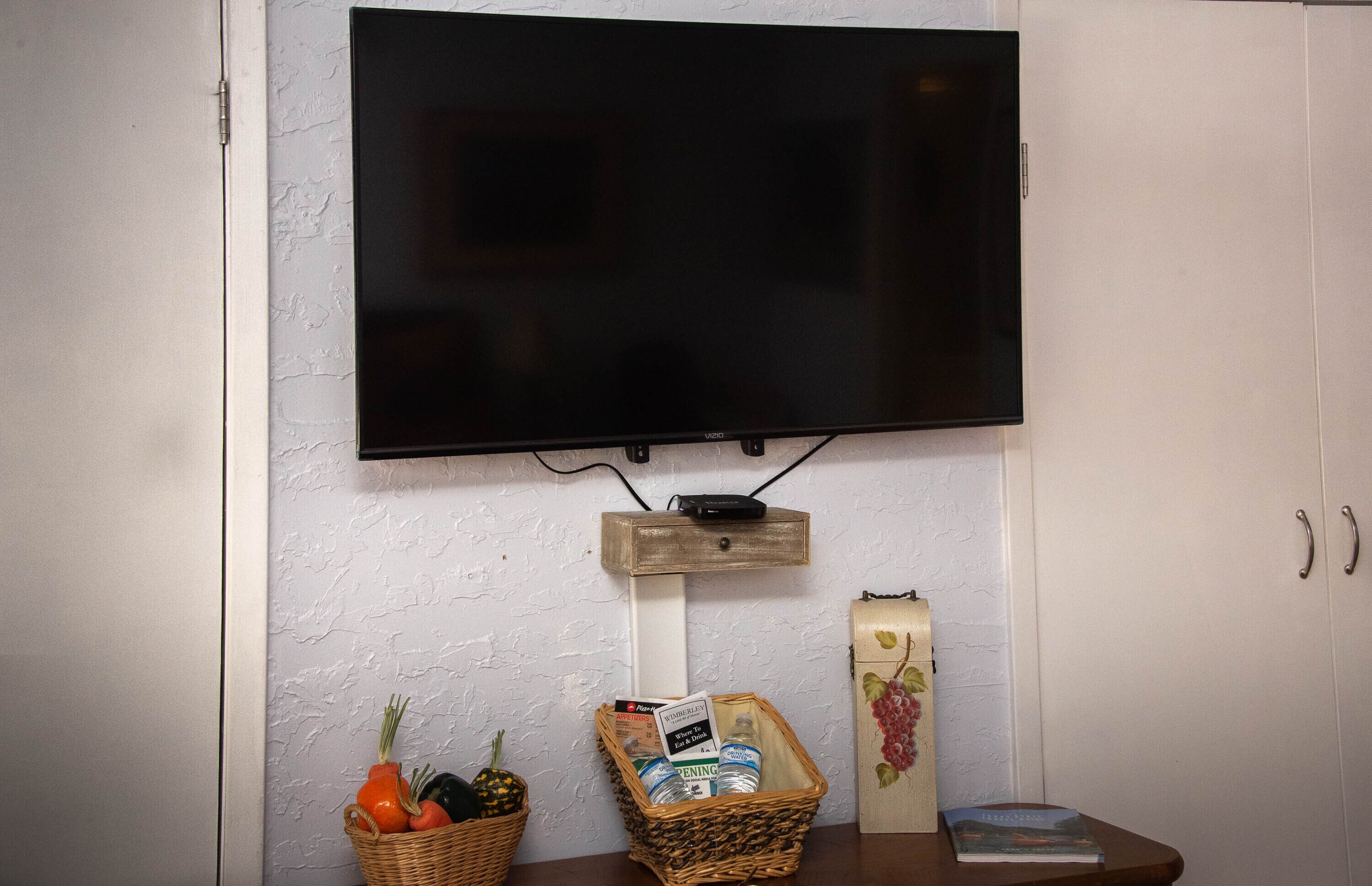 A flat-screen TV mounted on a textured wall, a wooden shelf below it, a wicker basket with vegetables, and a book on a table.