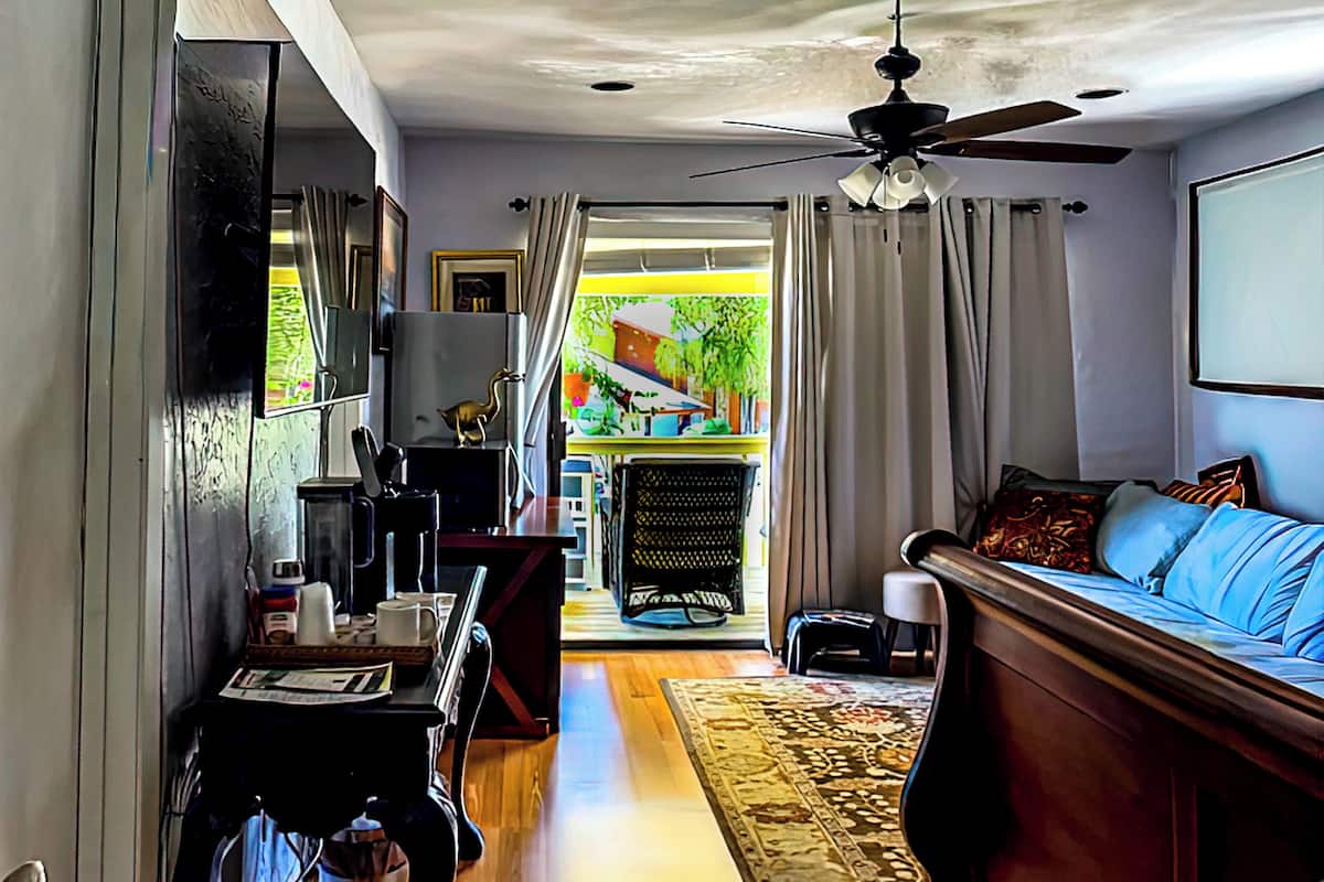 Een kamer met een houten vloer, een bed met blauw beddengoed, een bureau met een computer, een stoel en een raam met gordijnen.