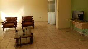 Un salon avec deux fauteuils en bois, une table basse en verre, un téléviseur posé sur un meuble en bois et une porte-fenêtre coulissante donnant sur la cuisine.