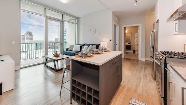 A modern kitchen with a central island, a dining area with a table and chairs, and a view of the city through large windows. - Heaven in Charlotte Furnished Apartments (Charlotte)