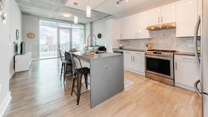 A modern kitchen with white cabinets, a central island, stainless steel appliances, and a large window overlooking a cityscape. - Heaven in Charlotte Furnished Apartments (Charlotte)