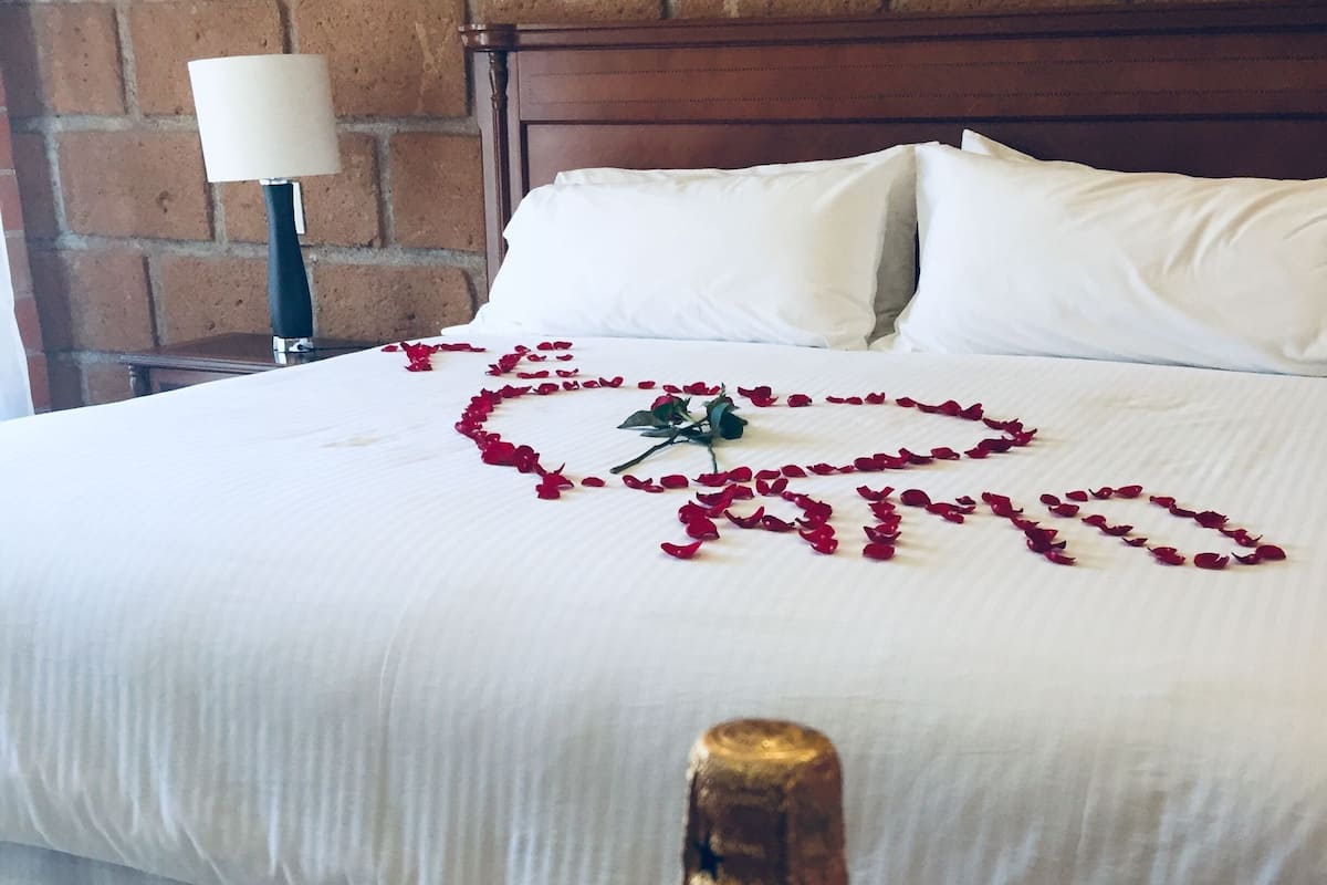 A bed with white linens, a heart-shaped arrangement of rose petals, and a bottle of champagne in front of it.
