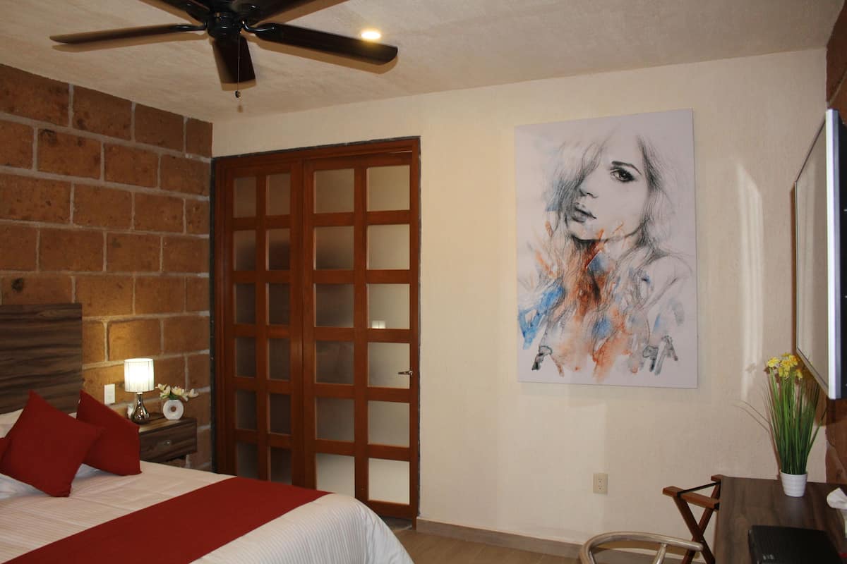 A bedroom with a bed, a wooden door, a ceiling fan, a painting, and a desk with a vase of flowers.