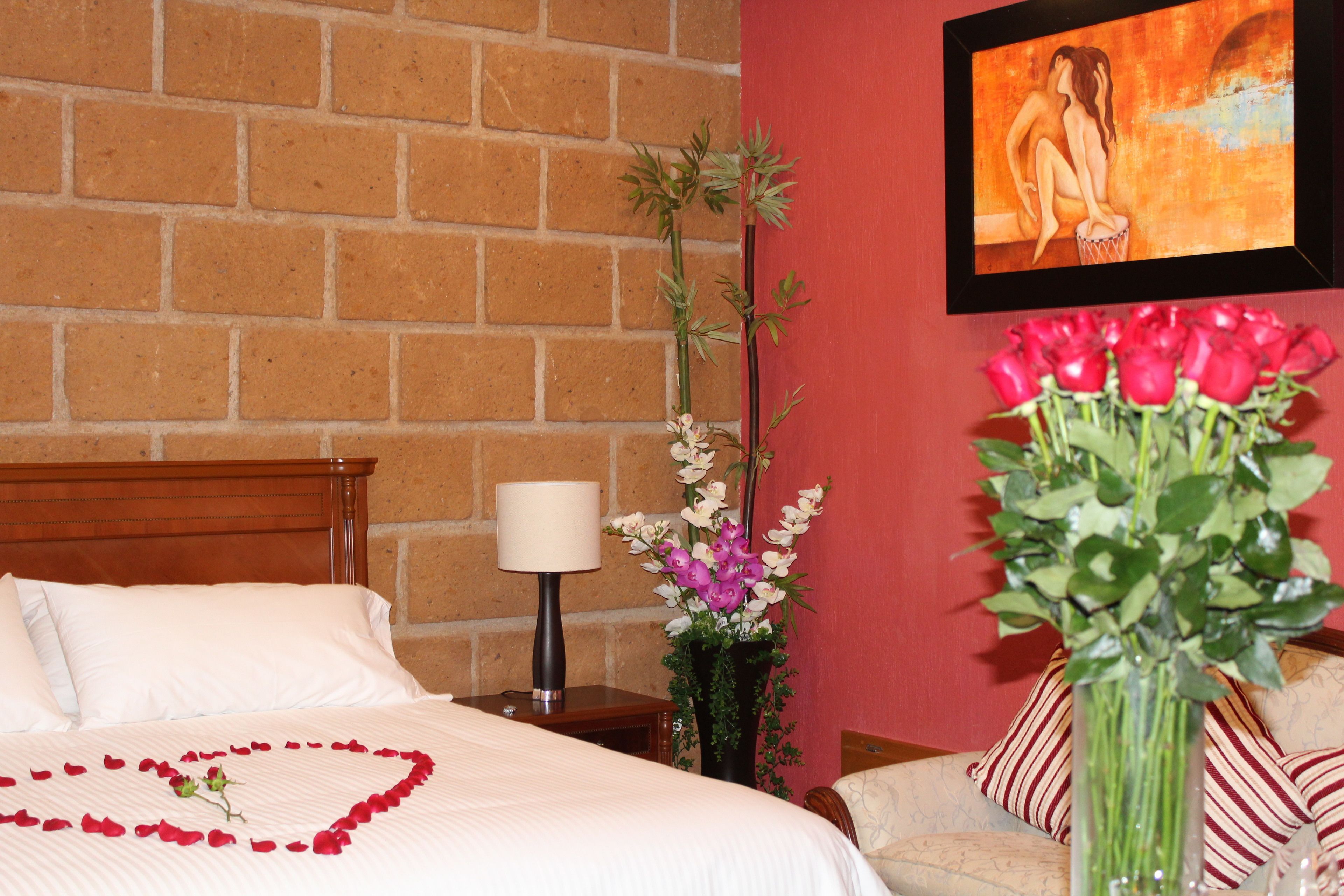Un cuarto de hotel con pared de ladrillo, una cama con sábanas blancas, una mesita de noche con lámpara, un florero con flores y un cuadro en la pared.