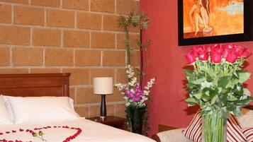 Un cuarto de hotel con pared de ladrillo, una cama con sábanas blancas, una mesita de noche con lámpara, un florero con flores y un cuadro en la pared.