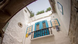 Un escalier menant à une porte rouge ornée d’une fenêtre, un balcon avec des volets bleus et un sol recouvert de carreaux de pierre.