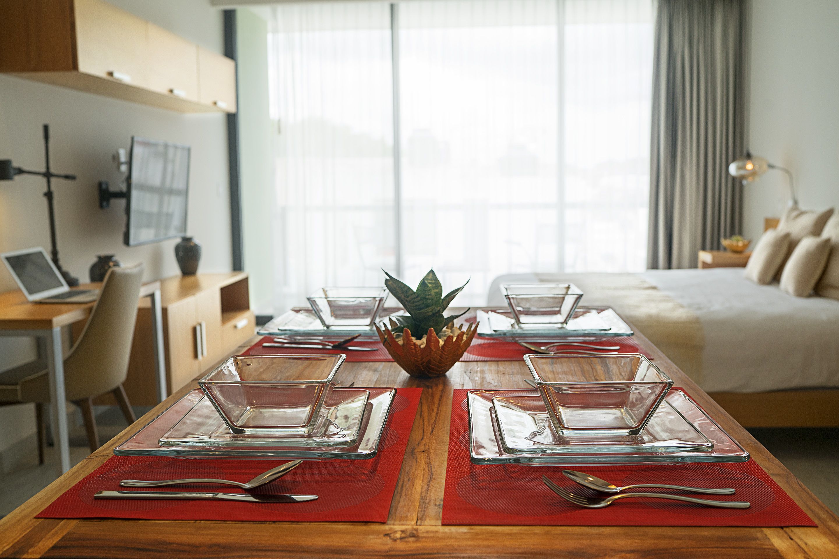 A neatly arranged dining table with clear glassware and red placemats, a laptop on a desk, and a bed in the background.