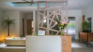 A modern reception desk with a laptop, a decorative wooden partition, and a potted plant with flowers. - Opal Apt. 202 Top Host Service, Ideal Location, Great Amenities (Playa del Carmen)