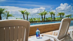 A poolside area with white chairs and a table, a bottle of water, and a view of palm trees and a clear sky. - Opal Apt. 202 Top Host Service, Ideal Location, Great Amenities (Playa del Carmen)