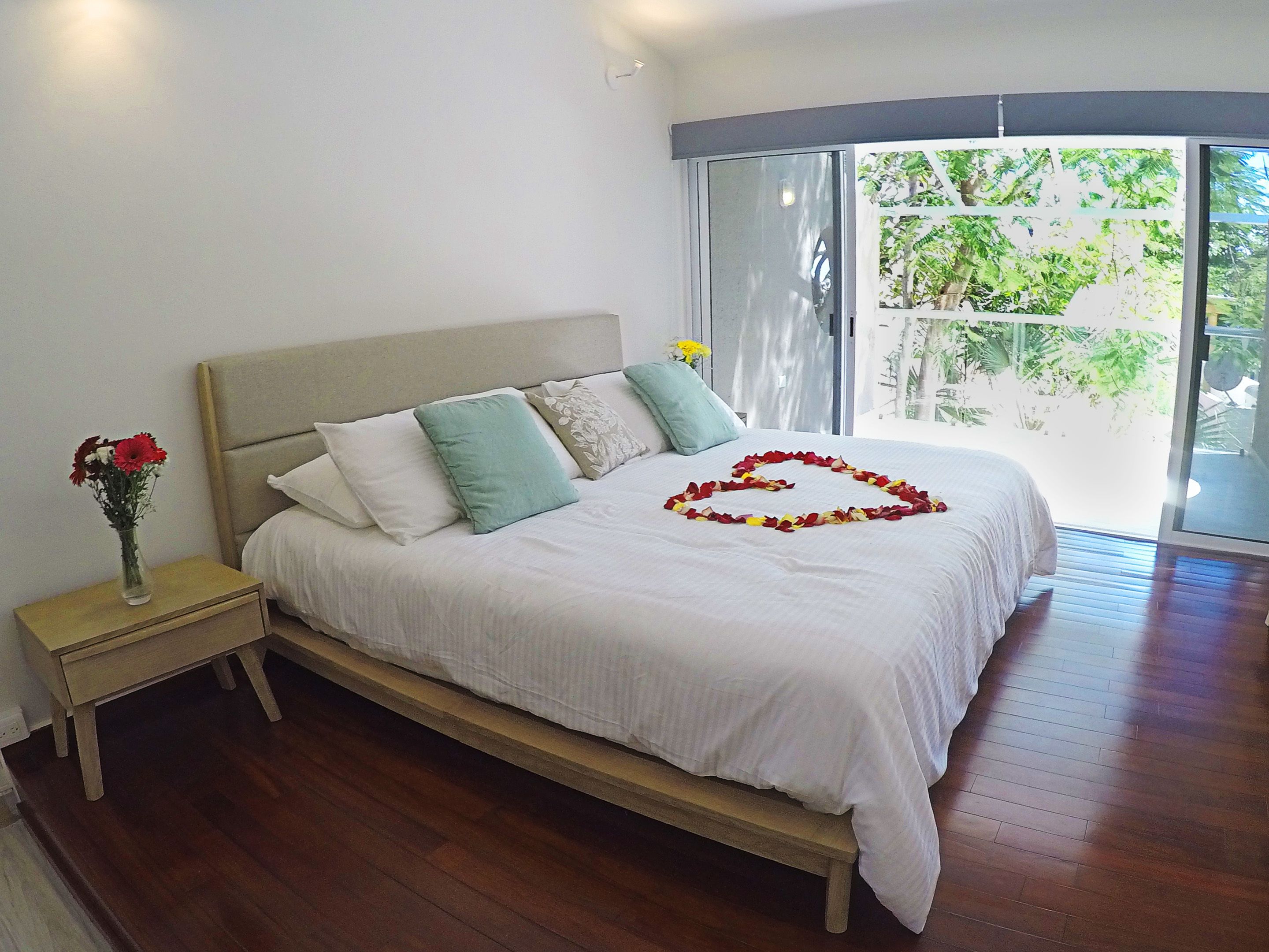 A neatly made bed with a heart-shaped flower arrangement, a vase with flowers, and a view of greenery through a large window.
