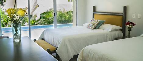 Quarto de hotel com duas camas, uma mesa de jantar e vista para uma piscina e áreas verdes lá fora.