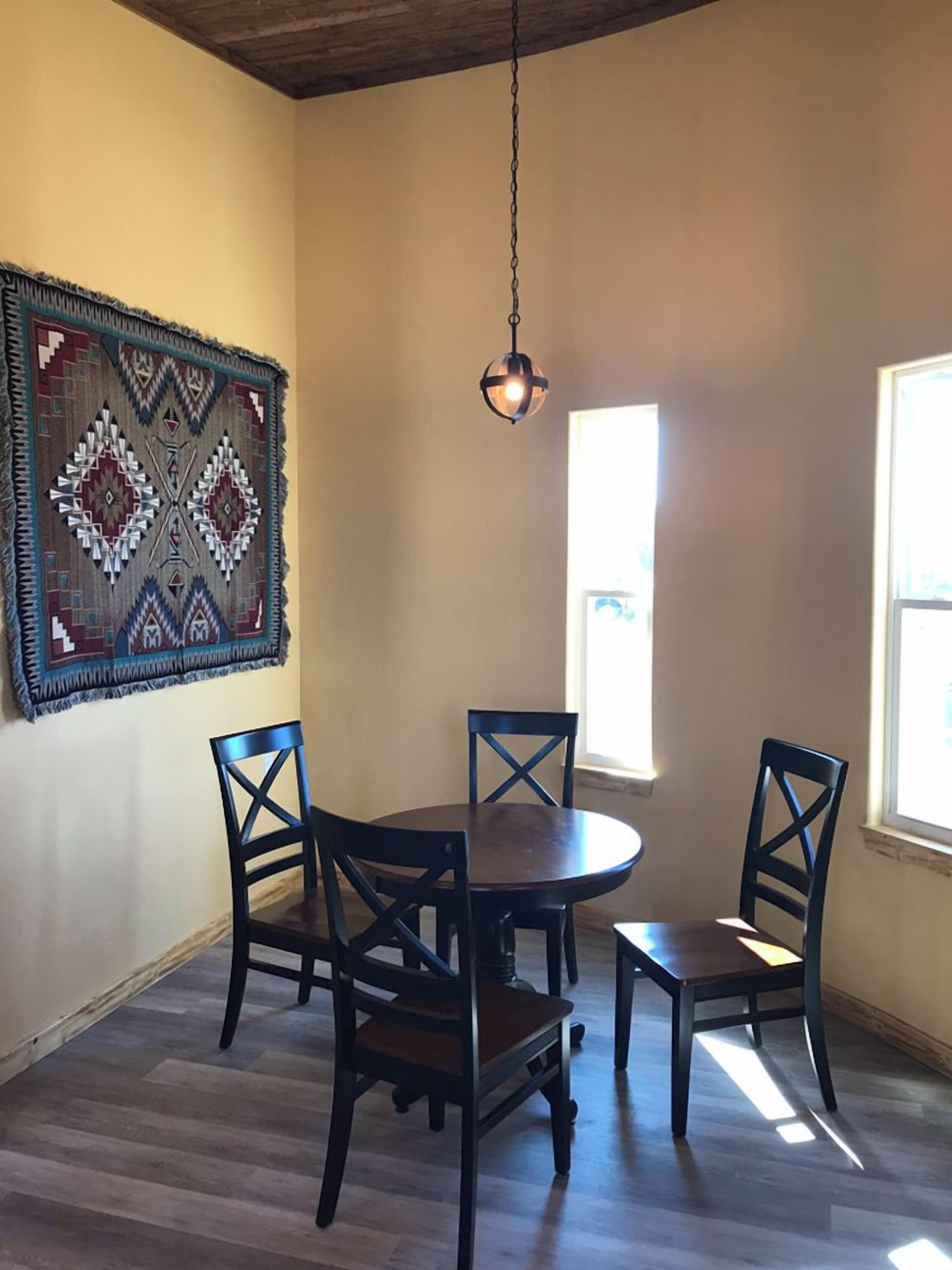A dining area with a round table and four chairs, a hanging light fixture, and a large patterned rug on the wall.