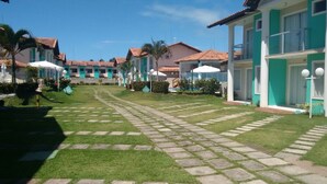 A courtyard with a paved walkway, surrounded by buildings with green and white facades, palm trees, and outdoor umbrellas. - Casa 13 Condomínio Mont Moriá (Porto Seguro)