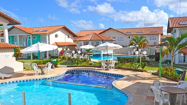 A swimming pool surrounded by outdoor seating, with houses in the background. - Casa 13 Condomínio Mont Moriá (Porto Seguro)