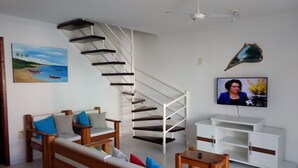 A living room with a wooden sofa, a small wooden table, a white TV stand, a white cabinet, a white staircase, and a ceiling fan. - Casa 13 Condomínio Mont Moriá (Porto Seguro)
