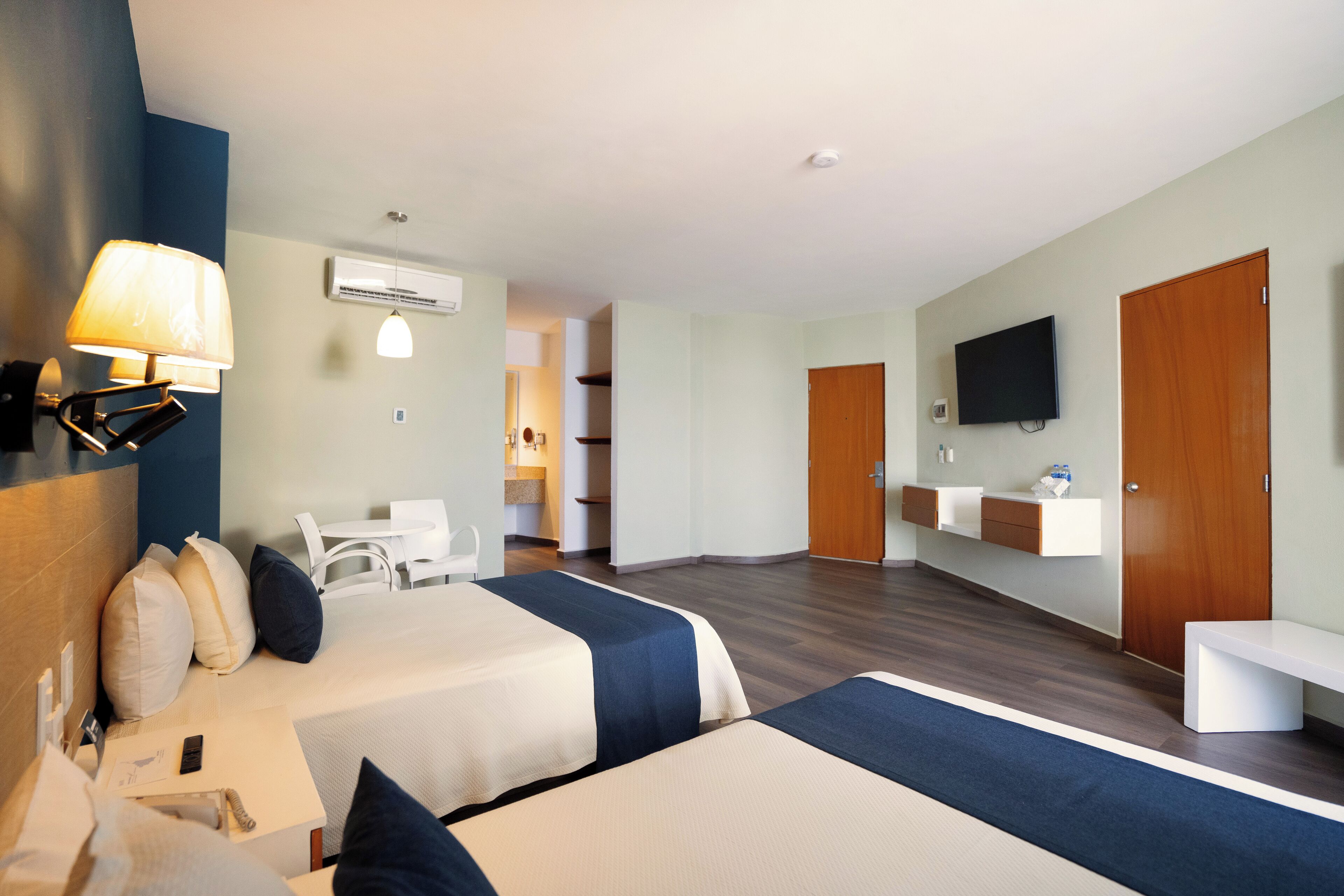 A hotel room with two beds, a TV, a desk, a chair, and a bathroom visible in the background.