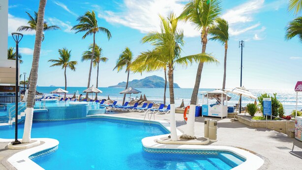 A pool area with blue lounge chairs and palm trees, surrounded by a clear sky and a view of the ocean. - Oceano Palace Beach Resort (Mazatlán)