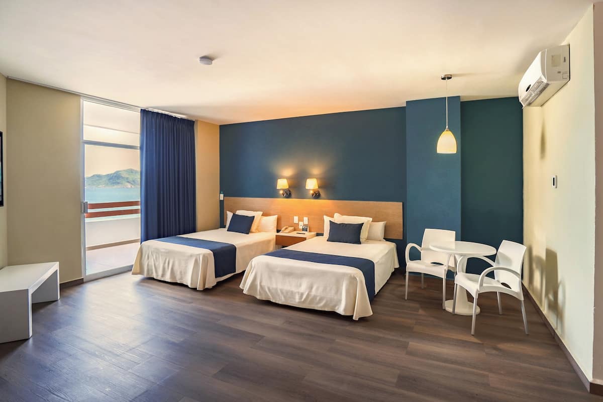 A hotel room with two beds, a desk, a chair, and a view of the sea.
