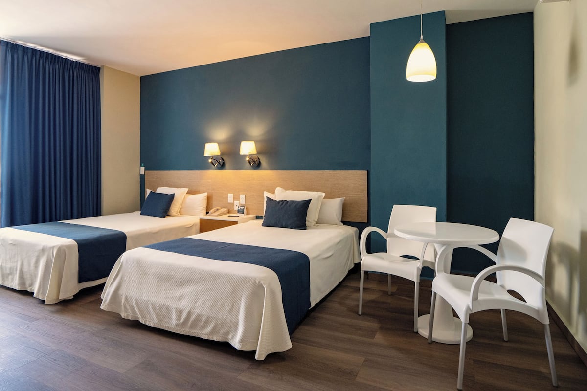 A hotel room with two beds, a small table with chairs, a desk with a lamp, and a hanging light fixture.