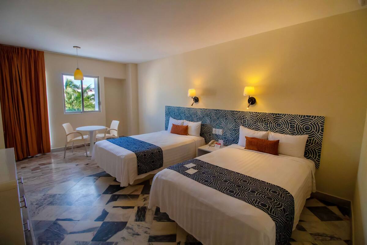 A hotel room with two beds, a small table, a chair, a window with curtains, and a patterned headboard.