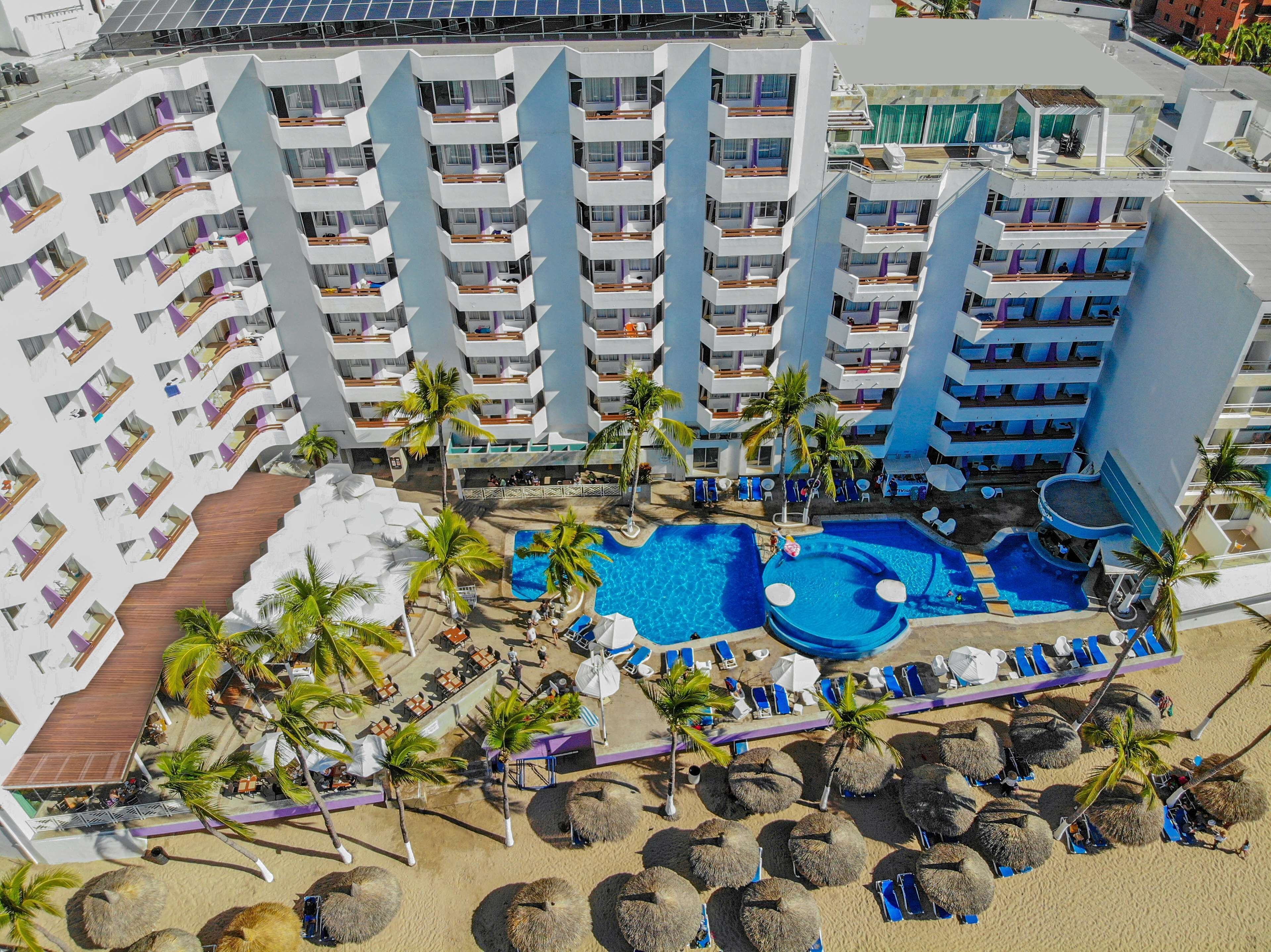 Un complexe hôtelier doté de nombreux balcons, d’une piscine centrale et d’une plage aménagée avec des chaises longues et des palmiers.