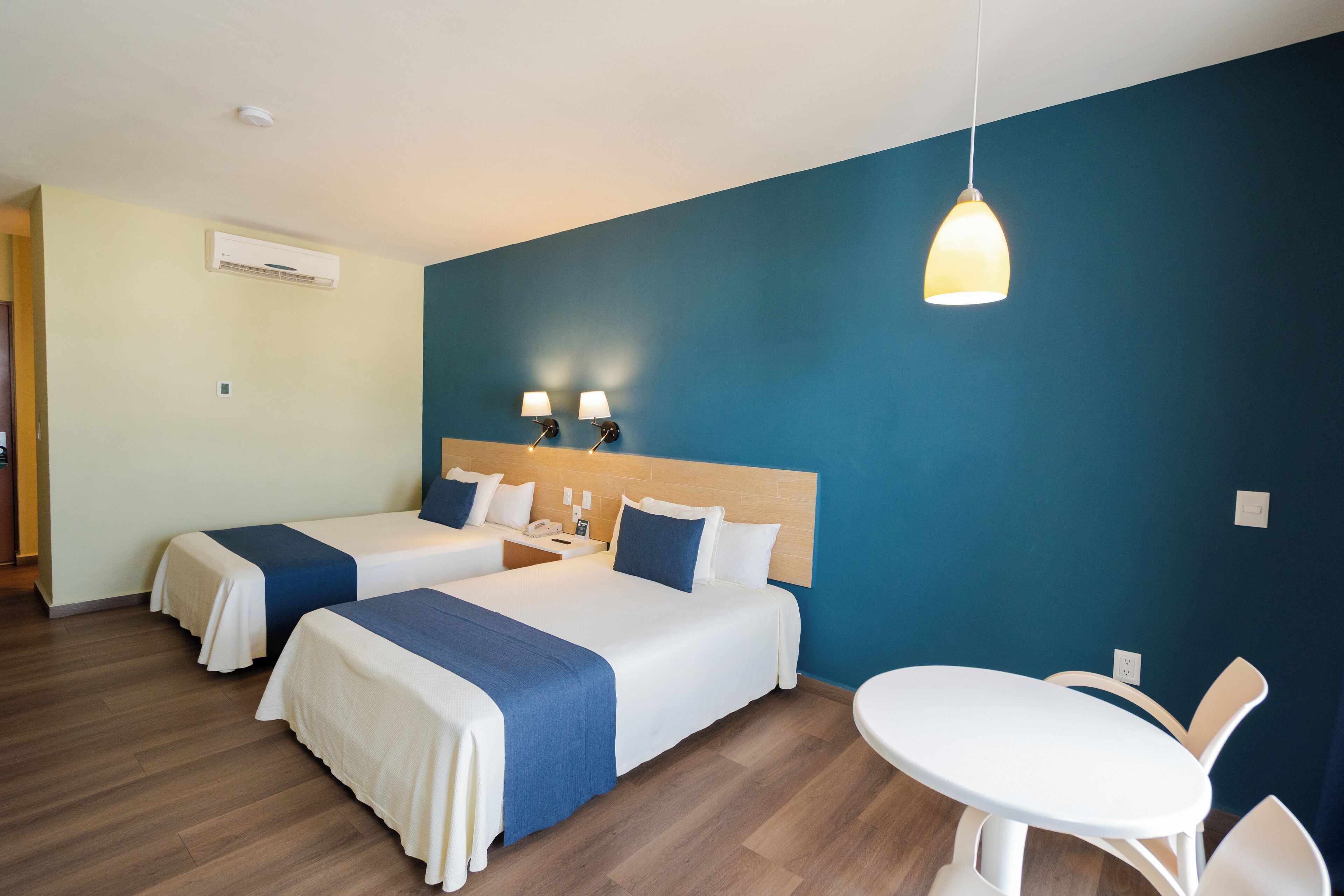 Habitació d'hotel amb dos llits, una taula petita i una cadira. Hi ha una làmpada al sostre i un aparell d'aire condicionat a la paret.
