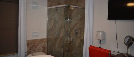 A hotel room with a bed, a shower with glass doors, a red chair, a desk with a lamp, and a TV on the wall.