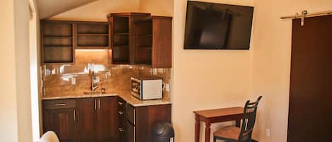 A compact kitchen with dark wood cabinets, a granite countertop, and a built-in microwave. There is a small dining table with a chair, a trash can, and a flat-screen TV mounted on the wall.
