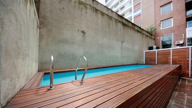 A wooden deck with a swimming pool, a concrete wall, and a building with windows in the background.