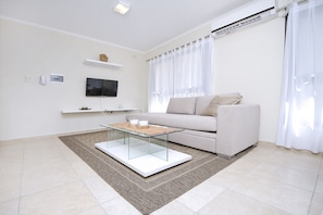A modern living room with a glass coffee table, a sofa, a flat-screen TV, and a white air conditioning unit.