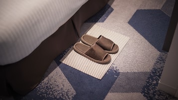 Braune Hausschuhe auf einer strukturierten Matte neben einem Bett in einem Hotelzimmer.