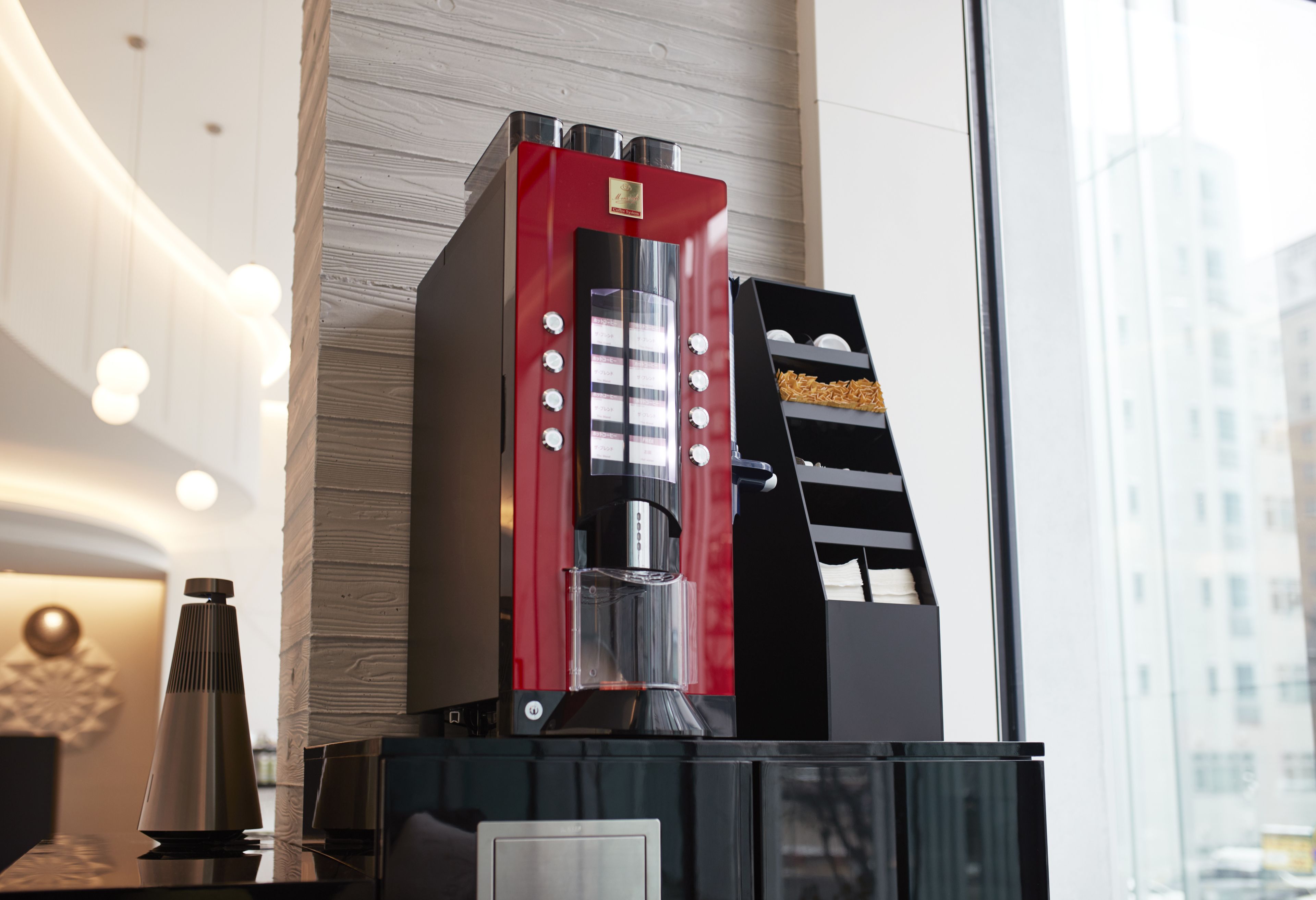 A modern coffee machine with a red and black design, placed on a black counter next to a window with a view of buildings.