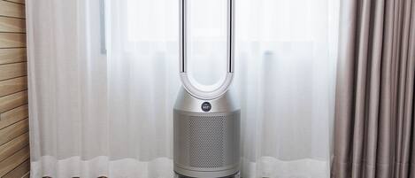 Un ventilador sin aspas Dyson en una habitación con piso de madera y una ventana con cortinas de gasa.