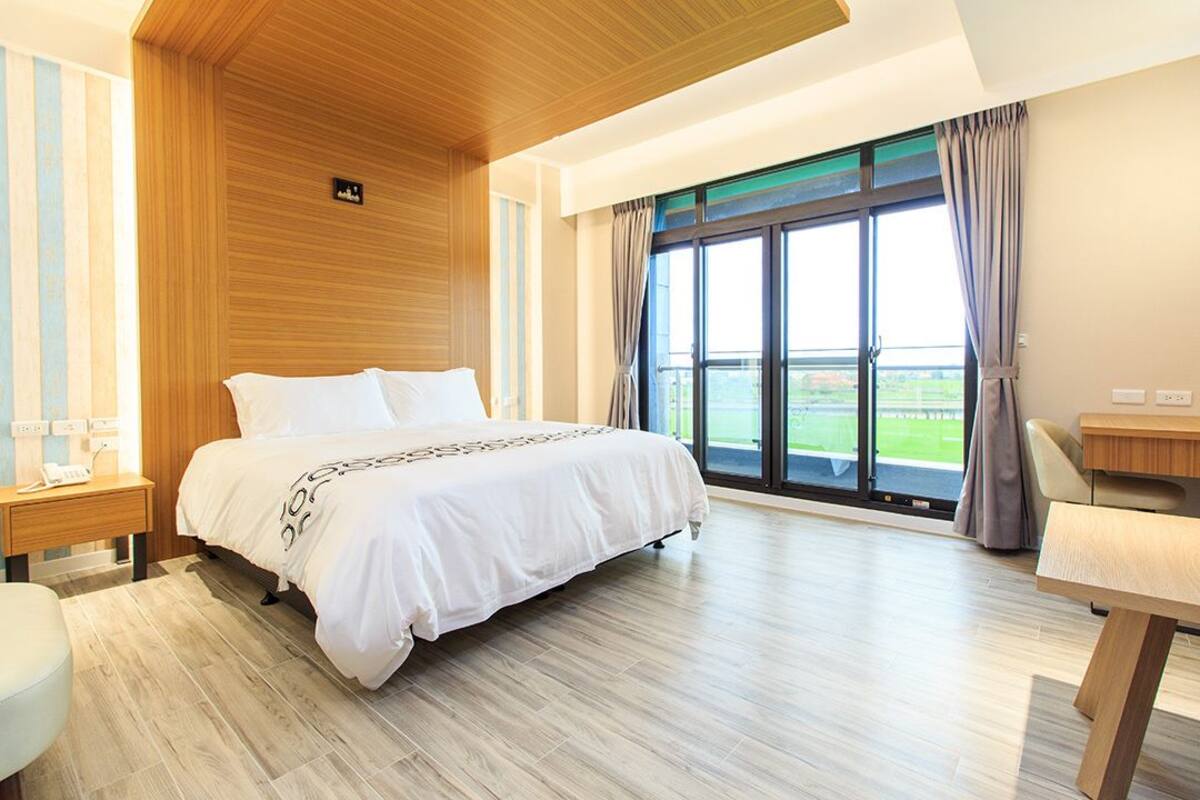 A modern hotel room with a large bed, wooden ceiling, and a view of a green landscape through a large window.