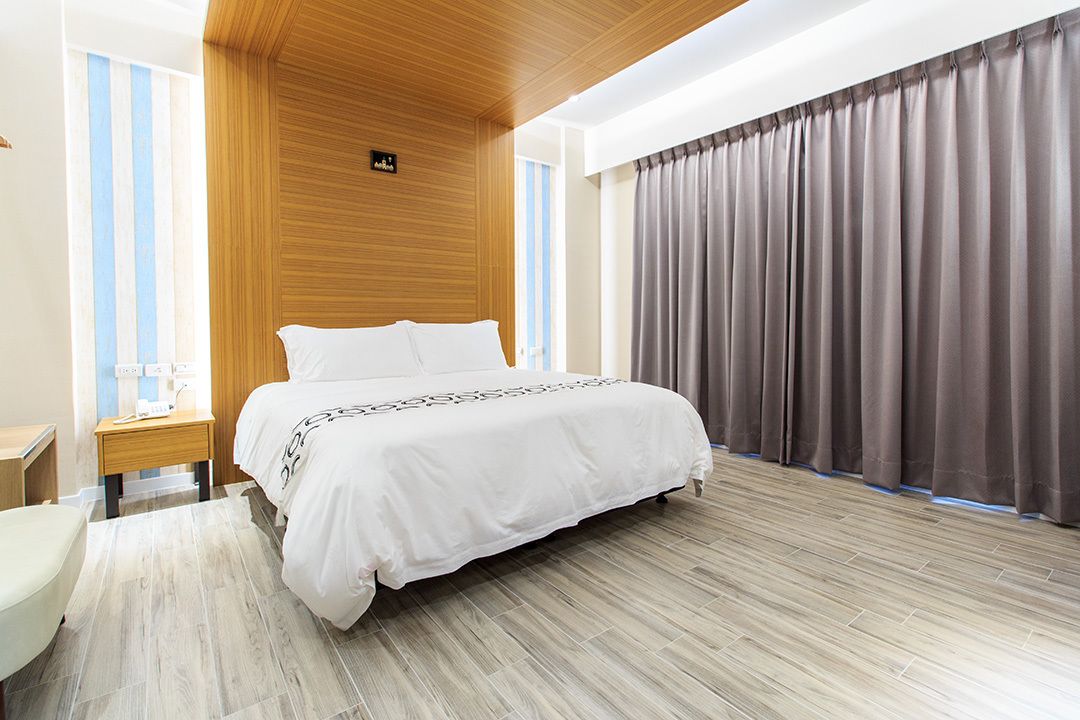 A modern hotel room with a large bed, wooden paneling on the walls, a small bedside table, and a view of the city through large windows.