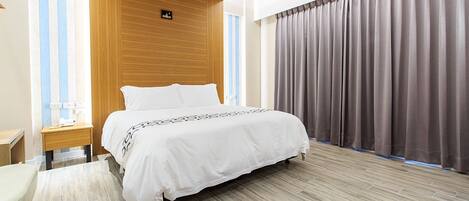 A modern hotel room with a large bed, wooden paneling on the walls, a small bedside table, and a view of the city through large windows.