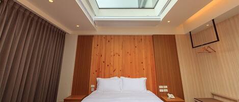Habitación de hotel con tragaluz, paredes de paneles de madera, una cama con ropa blanca, una mesita de noche con teléfono y un escritorio.