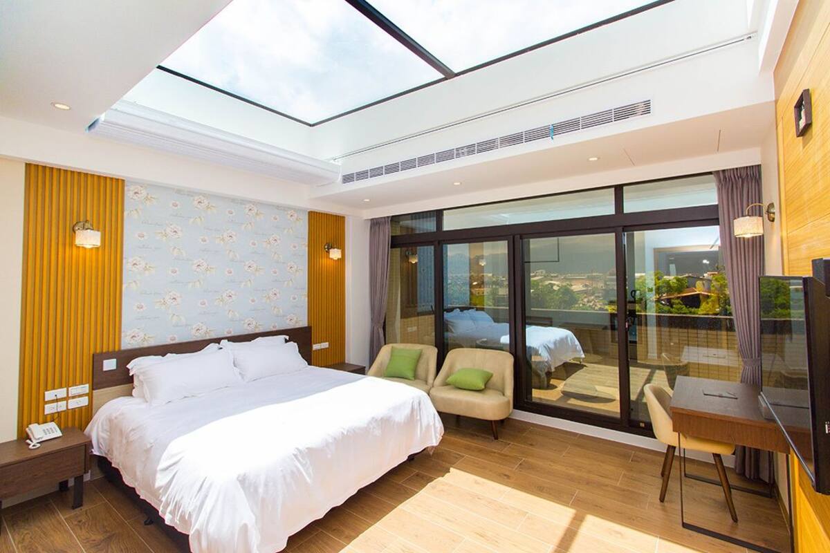 A modern hotel room with a large bed, two armchairs, a desk, and a large window offering a city view.
