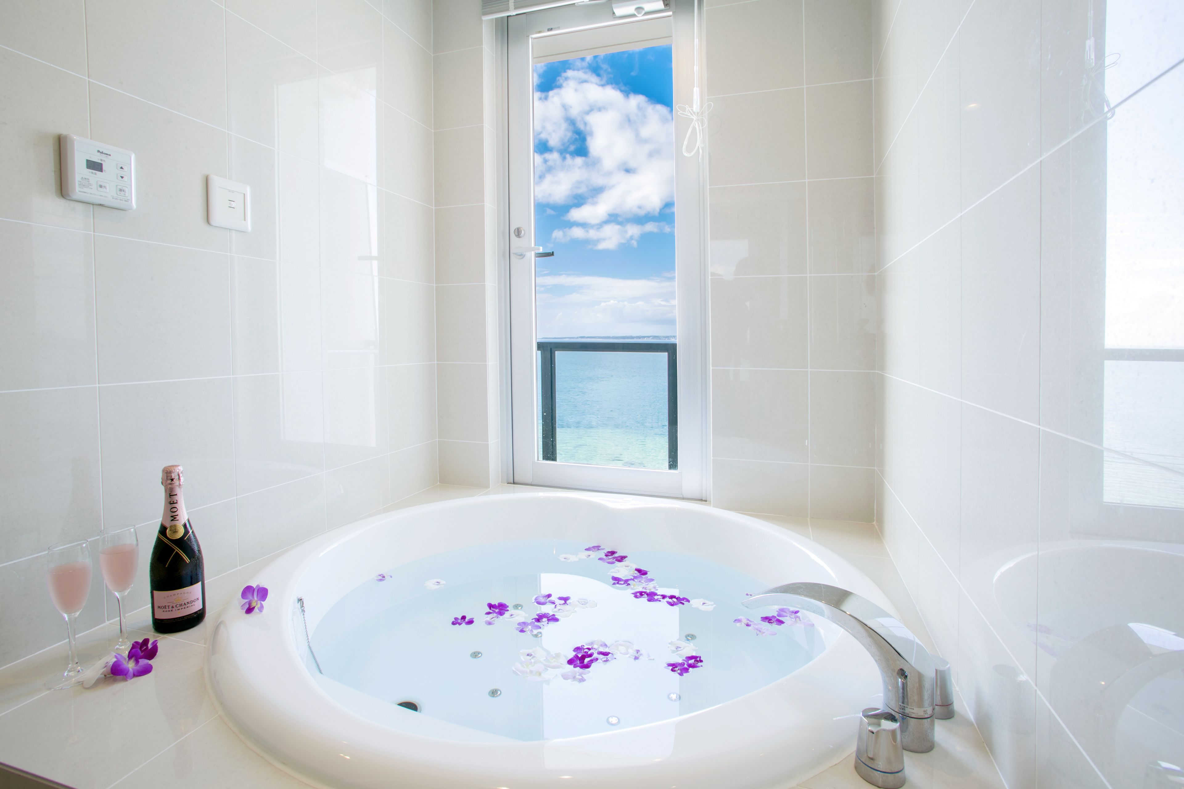 A bathroom with a large bathtub filled with water and purple flowers, a bottle of champagne, and two glasses on the counter.