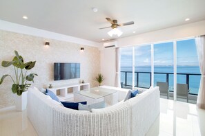 A modern living room with a white wicker sofa, a glass coffee table, and a flat-screen TV. There is a large window offering a view of the ocean. - Costa Bella Condominiun Resort (Motobu)