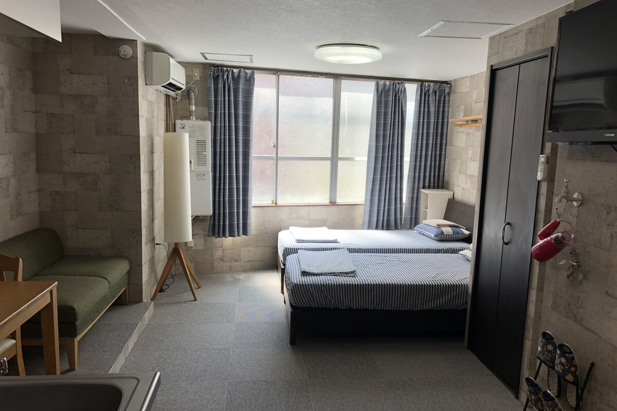 A compact hotel room with a bed, a sofa, a desk, a sink, and a window with curtains.