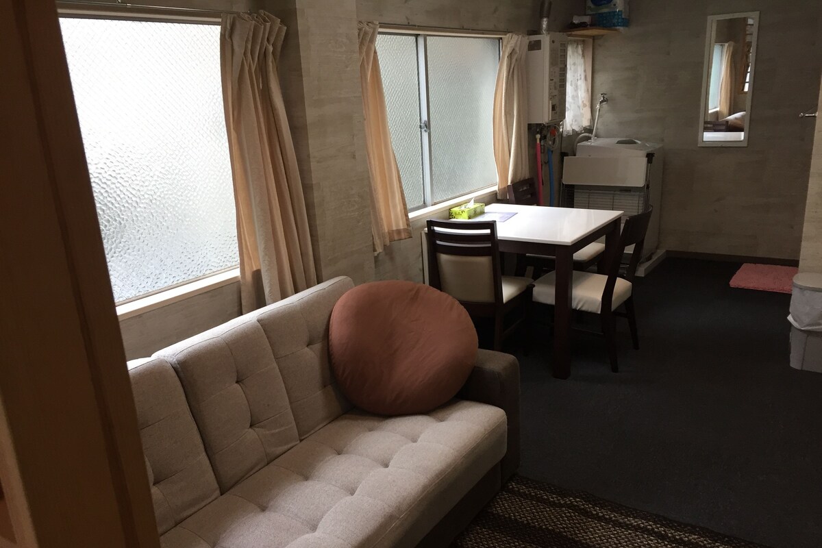 A compact living space with a sofa, a dining table, chairs, a small kitchen area with a sink and mirror, and a window with curtains.