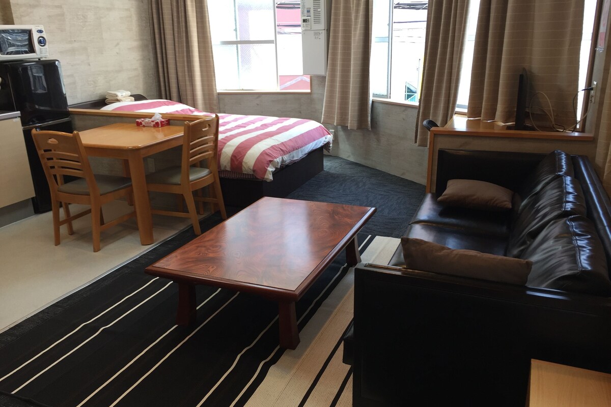 A compact hotel room with a bed, a wooden table, a black leather sofa, a small desk with a computer, and a microwave.