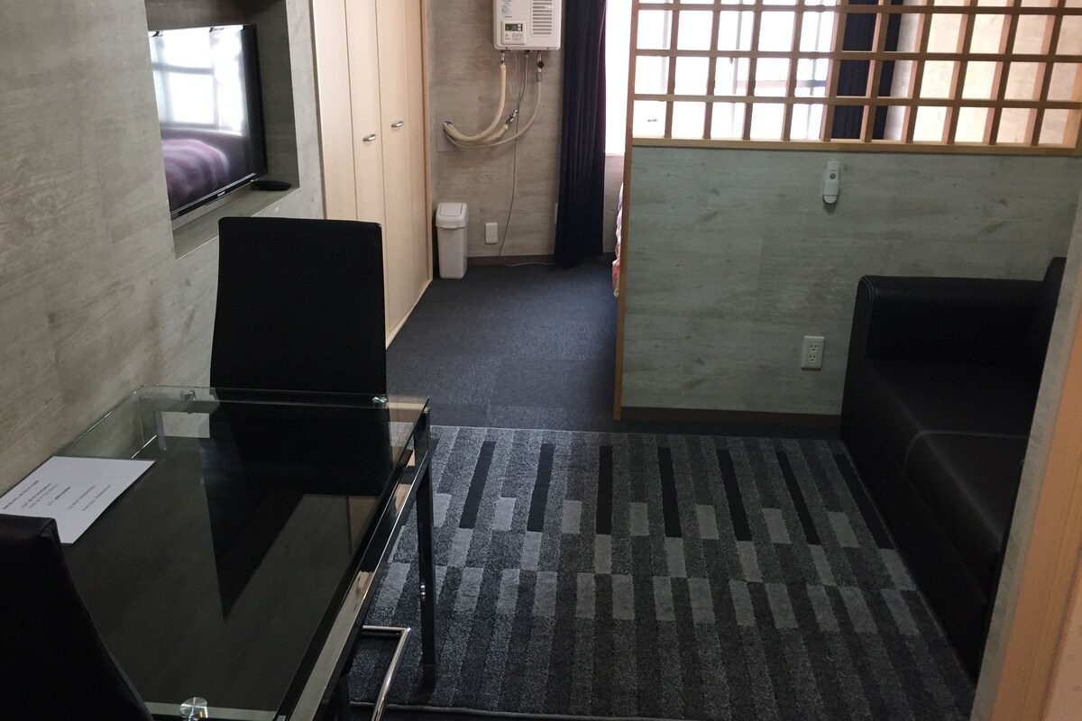A small, compact room with a bed, a desk, and a TV. There is a window with a view of the outside, and a heater mounted on the wall.