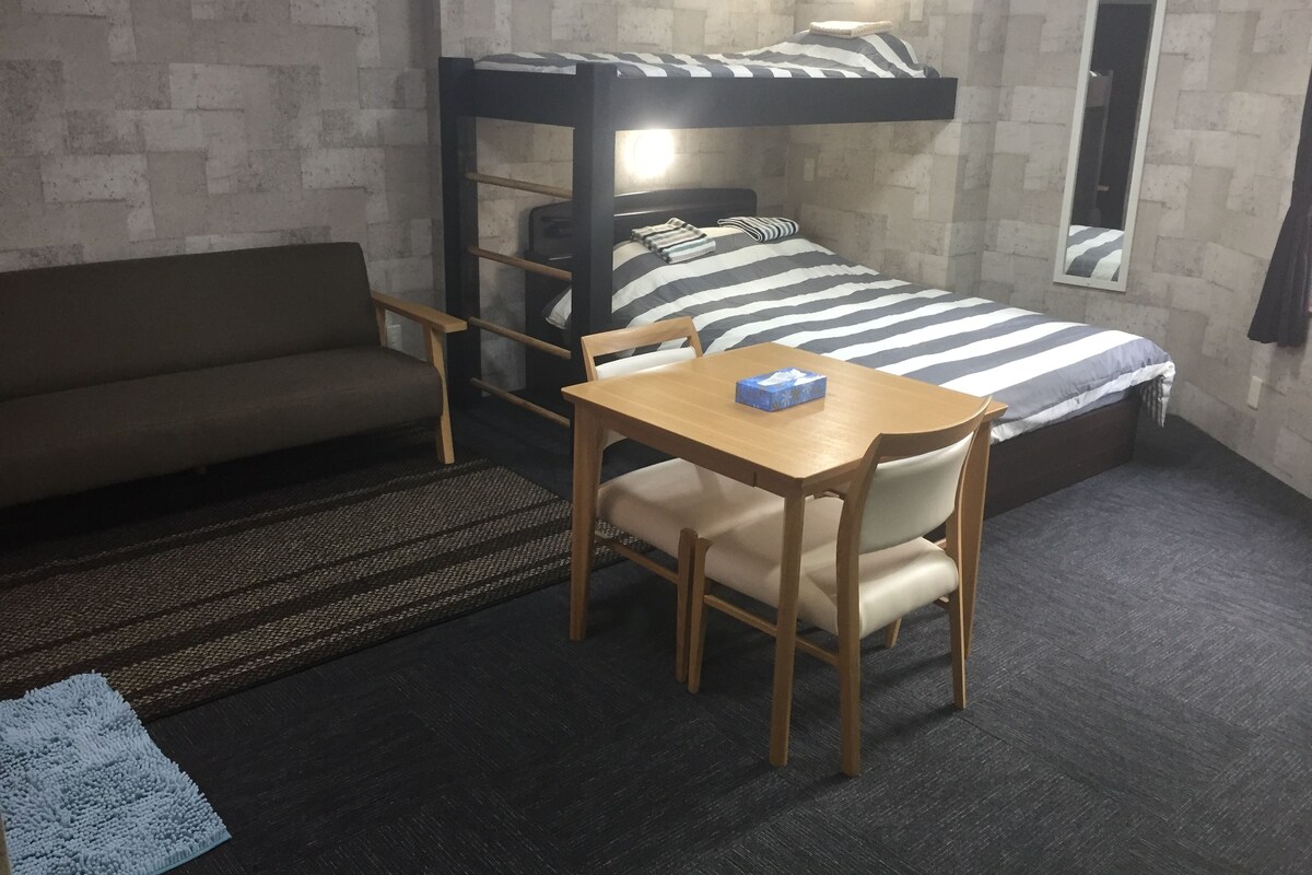 A dorm-style room with a bunk bed, a sofa, a table, and a chair.