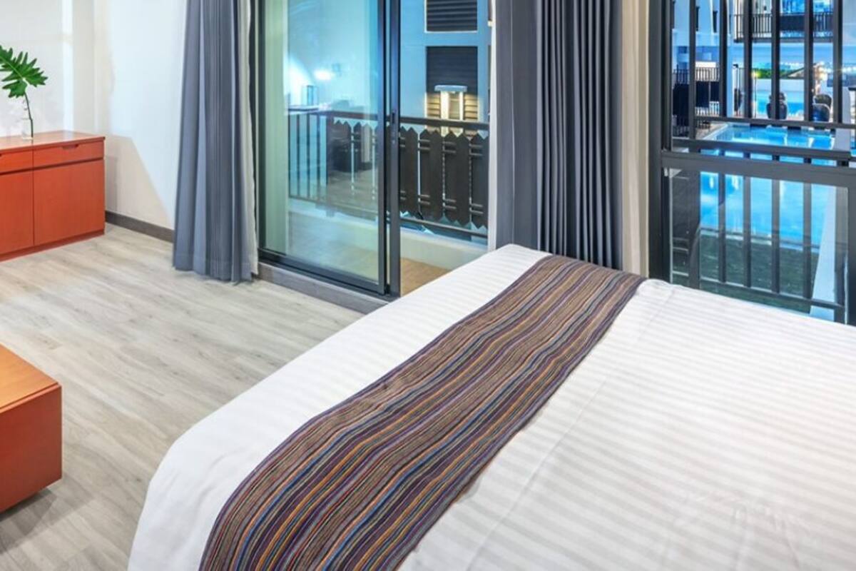 Quarto de hotel com cama, escrivaninha com televisão, cadeira, mesa de centro com planta, e vista para uma piscina e varanda.
